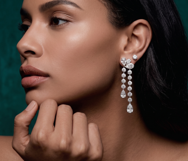 A close-up lifestyle shot of a model wearing a dramatic, multi-tiered dangle earring set with clear gemstones on a dark green background.
