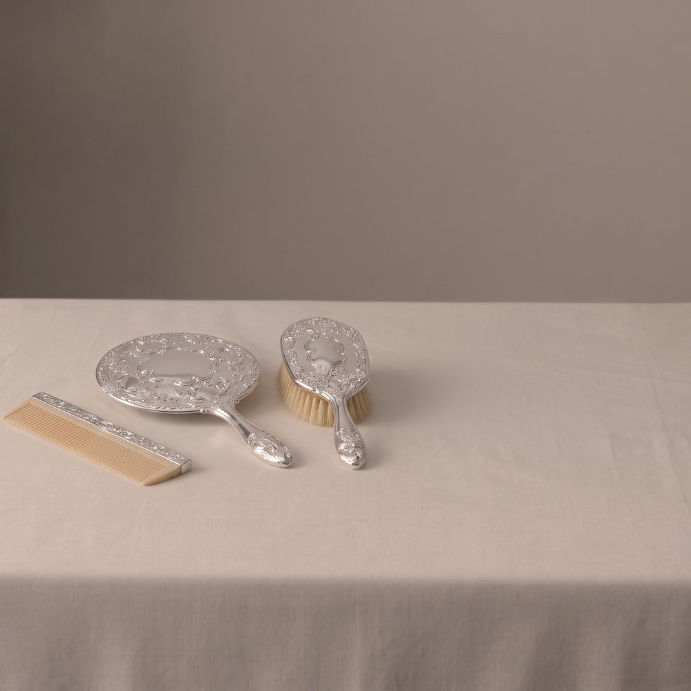Ornate silver engraved hand mirror, hairbrush, and ivory-colored comb on a white cloth.