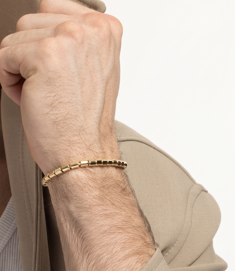 A close-up of a man's wrist wearing a thin yellow gold beaded bracelet, dressed in a beige blazer over a striped shirt.