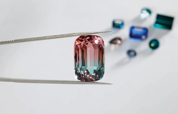 Emerald cut bi-color pink and teal gemstone held by tweezers with loose colored gems.