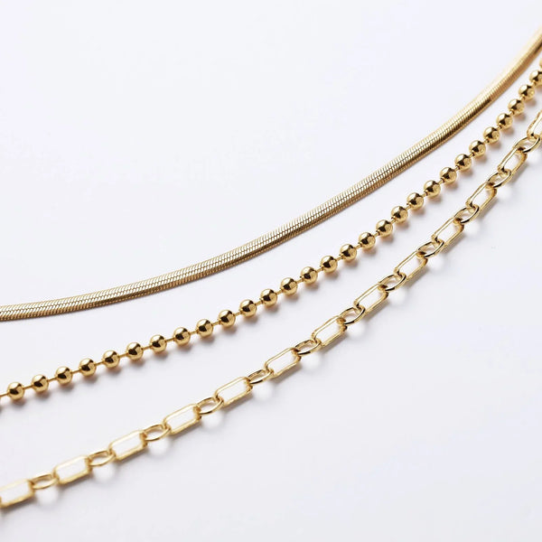 Three layered gold necklaces featuring snake, bead, and paperclip chain styles.