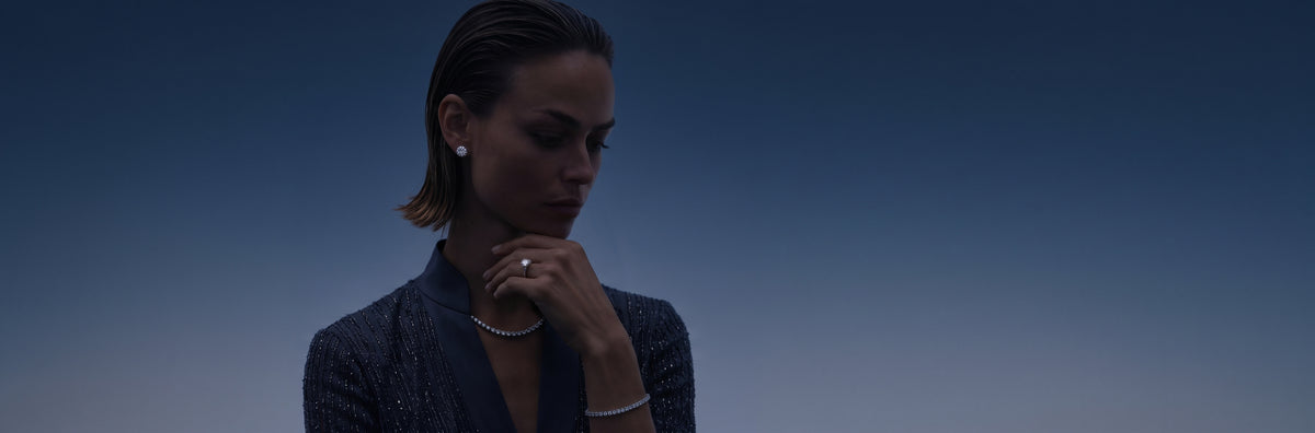A woman in a dark textured blazer wearing delicate jewelry, including a ring, earrings, and bracelet, set against a dark blue gradient background.