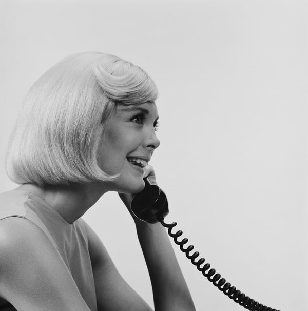 Black and white vintage photo of a smiling woman with a blonde bob talking on a rotary phone.