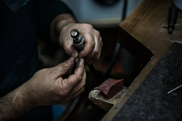 Jeweler's hands using a rotary tool to craft fine jewelry at a professional workbench.