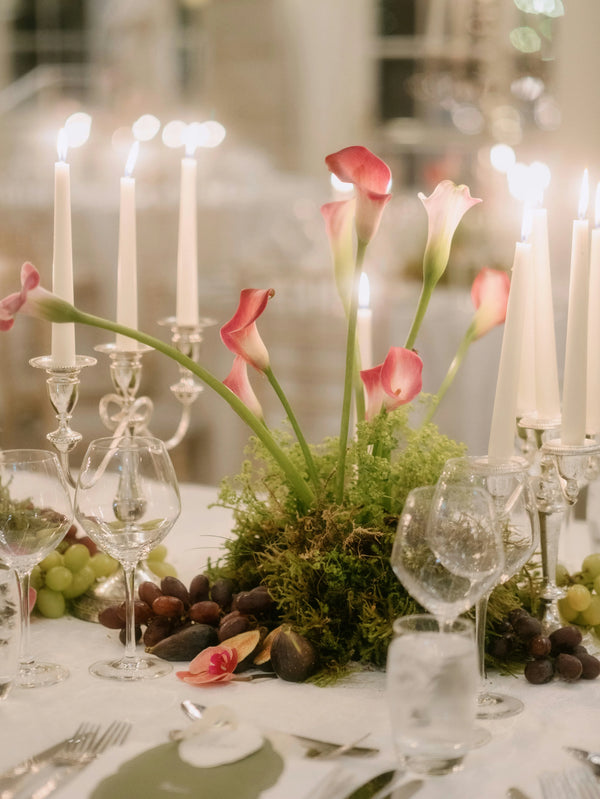 Elegant wedding table centerpiece with pink calla lilies, moss, and lit white taper candles.