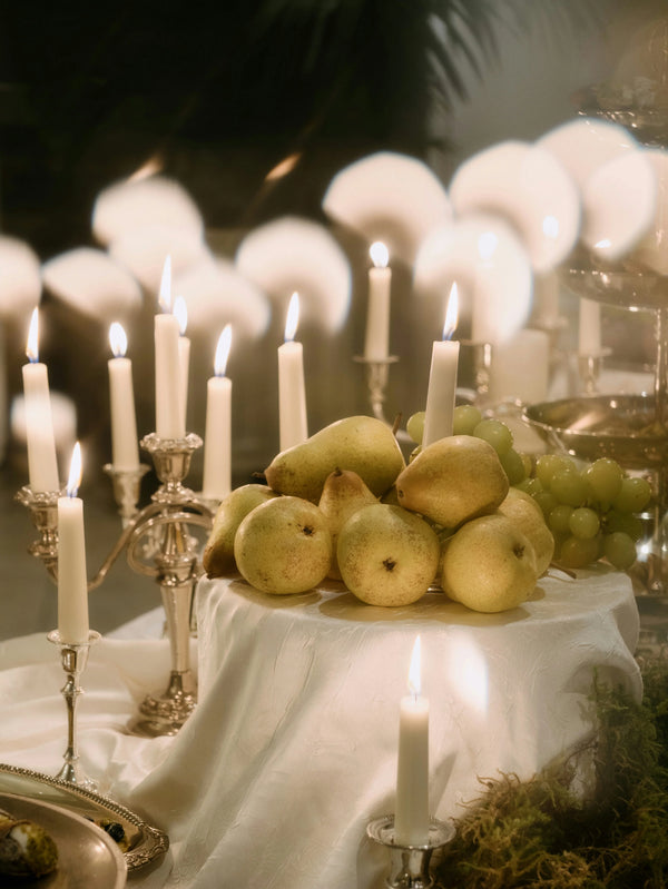 Still life of fresh pears and green grapes arranged with glowing white candles on a white cloth.